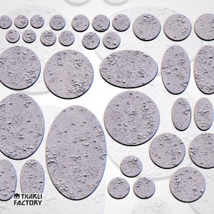 May include: A collection of various circular and oval-shaped grey bases with a textured, rocky, and cracked earth surface. The bases are arranged in different sizes, and the logo Txarli Factory is printed in the bottom left corner.