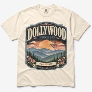 May include: A cream-colored short-sleeve t-shirt featuring a graphic print of a sunset over mountains with the text DOLLYWOOD TENNESSEE and SMOKY MOUNTAIN MEMORIES. The design includes a butterfly, pink flowers, and a heart icon within a decorative frame.