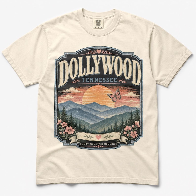 May include: A cream-colored short-sleeve t-shirt featuring a graphic print of a sunset over mountains with the text DOLLYWOOD TENNESSEE and SMOKY MOUNTAIN MEMORIES. The design includes a butterfly, pink flowers, and a heart icon within a decorative frame.