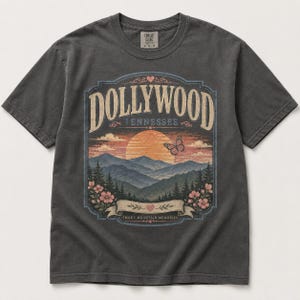 May include: A charcoal gray short-sleeve t-shirt featuring a graphic print of a sunset over mountains with the text DOLLYWOOD TENNESSEE and SMOKY MOUNTAIN MEMORIES. The design includes a butterfly, flowers, and hearts in muted orange, pink, and blue.
