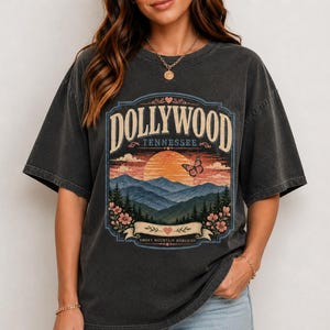 May include: A charcoal gray short-sleeve t-shirt featuring a graphic print of a mountain landscape at sunset. The text reads DOLLYWOOD TENNESSEE and SMOKY MOUNTAIN MEMORIES. The design includes a butterfly, flowers, and hearts in a vintage style.