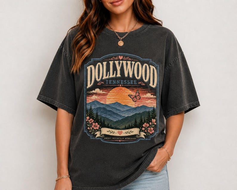 May include: A charcoal gray short-sleeve t-shirt featuring a graphic print of a mountain landscape at sunset. The text reads DOLLYWOOD TENNESSEE and SMOKY MOUNTAIN MEMORIES. The design includes a butterfly, flowers, and hearts in a vintage style.