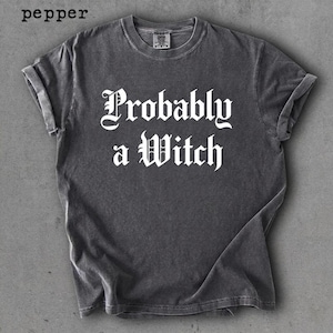 May include: A charcoal grey t-shirt with rolled sleeves featuring the white text 'Probably a Witch' in a gothic font. The shirt has a washed, vintage-style finish and a crew neckline. The word 'pepper' is printed in the top left corner.