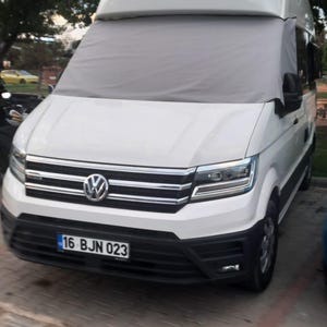 Op de afbeelding: Een witte Volkswagen Crafter camper met een hoog dak en een grijs privacyscherm over de voorruit. Het voertuig is voorzien van een zwarte voorbumper, chromen grille-accenten en een kentekenplaat met de tekst 16 BJN 023.