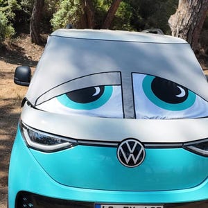 May include: A light blue and white Volkswagen electric vehicle featuring a windscreen sunshade printed with a large, sleepy-looking cartoon eye design. The eyes are white with teal irises and black pupils, positioned to cover the entire front windscreen.