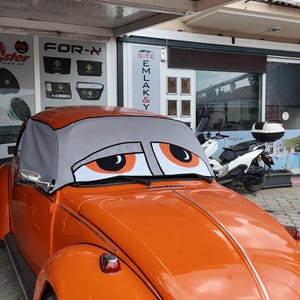 May include: A bright orange vintage car featuring a grey windscreen sunshade printed with large, sleepy-looking orange eyes. The car has a rounded bonnet, a chrome headlight, and a chrome side mirror. The windscreen cover displays a cartoonish expression.