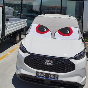 May include: A white Ford vehicle featuring a grey windscreen sunshade printed with a cartoon-style graphic of two large, angry-looking eyes with red irises and black pupils, positioned across the front glass to cover the entire windscreen area.
