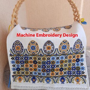 Könnte beinhalten: Weißes Stoff-Korbfutter mit detailliertem Maschinenstickerei-Design. Das Design zeigt bunte Ostereier und geometrische Muster in Blau, Gelb und Braun. Der Text "Machine Embroidery Design" ist sichtbar.