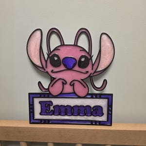 Peut inclure: Panneau décoratif rose et violet représentant un personnage avec le nom "Emma" en lettres pailletées violettes. Le personnage a de grandes oreilles, un nez violet et des yeux noirs. Le panneau est encadré d'une bordure violette.