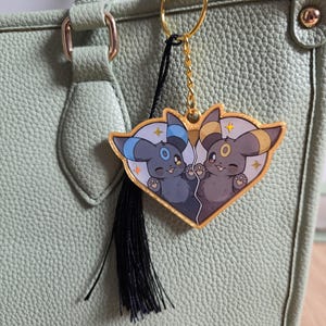 以下が含まれることがあります： 2つの漫画のキャラクターが描かれた、ハート型のチャームが付いたゴールドトーンのキーホルダー。 チャームは金の縁取りと黒いタッセルが付いています。 キーホルダーは、明るい緑色のテクスチャ加工されたハンドバッグに取り付けられています。