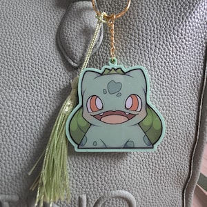 May include: A green Bulbasaur keychain with a gold-colored chain and keyring. The Bulbasaur design features a smiling face with orange eyes and a light green body. A green tassel hangs below the keychain. The keychain is attached to a light green handbag.