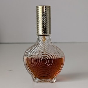 May include: A clear glass perfume bottle with a rounded, bulbous body featuring a swirling, embossed spiral pattern. The bottle contains a dark amber liquid and is topped with a cylindrical, metallic gold cap that has a diagonal ribbed texture.