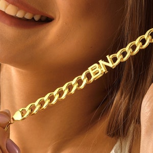 May include: A gold chain necklace with the word "BIND" spelled out in large letters. The necklace is worn by a person with long brown hair.