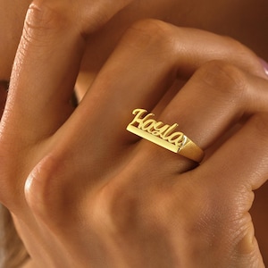 Op de afbeelding: Een goudkleurige ring met de naam "Hayla" in een cursief lettertype. De ring is een bandstijl, waarbij de naam de bovenkant van de band vormt. De ring wordt aan een vinger getoond.