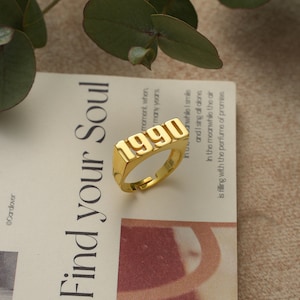 Könnte beinhalten: Goldring mit dem Jahr 1990 in Relief. Der Ring liegt auf einem weißen Hintergrund mit Text.