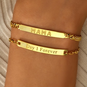 May include: Two gold bracelets are displayed. The top bracelet features the word "MAMA" engraved on a rectangular plate. The bottom bracelet has the words "Day 1 Forever" engraved on a similar plate. Both bracelets have a chain-link design.