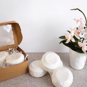 May include: A selection of white, round bath bombs, some wrapped in clear plastic, are presented with a brown cardboard box with a clear window. The box contains shredded paper. A white vase with a pink orchid is also in the image.