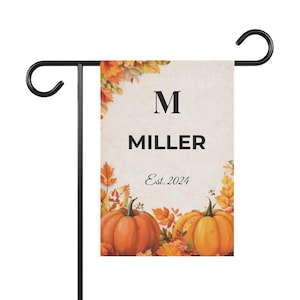 Könnte beinhalten: Ein personalisierter Gartenflagge mit dem Namen "MILLER" und dem Initial "M" in schwarzer Schrift. Der Hintergrund ist beige mit orangefarbenen Kürbissen und Herbstblättern. Der Text "Est. 2024" ist ebenfalls enthalten. Die Flagge ist auf einem schwarzen Metallständer.
