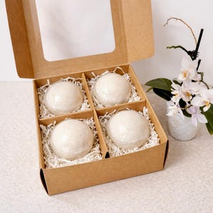 May include: A gift box with four bath bombs. Each bath bomb is round, light-colored, and encased in clear plastic. The bombs rest on white shredded paper inside a brown cardboard box with a clear window. A white orchid in a white pot is in the background.