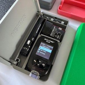 Op de afbeelding: Een grijze etui met een zwarte Accu-Chek Guide bloedglucosemeter, een zwarte Accu-Chek prikpen en een batterij. Het scherm van de meter toont tijd, datum en menu-opties. Een groene en roze container ernaast.