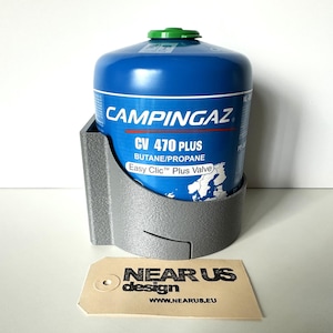 Könnte beinhalten: Eine blaue Campingaz CV 470 Plus Butan/Propan Gasflasche in einem grauen Halter. Die Flasche hat einen grünen Deckel und weißen Text. Der Halter ist aus grauem Kunststoff. Ein beigefarbenes Etikett mit dem Text "NEAR US design" befindet sich unten.
