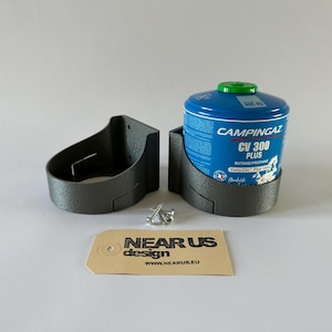 Könnte beinhalten: Eine blaue Campingaz CV 300 Plus Butan/Propan Gasflasche in einem dunkelgrauen Halter, mit einem passenden leeren Halter und einem kleinen Etikett mit der Aufschrift "NEAR US design". Die Flasche hat einen grünen Deckel.