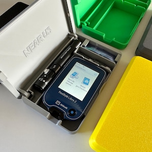 Può includere: Una custodia grigia con la scritta "NEARUS" stampata sul coperchio. La custodia è aperta e contiene un monitor di glucosio FreeStyle Libre 2 blu e bianco, una lancetta nera e argentata e un piccolo vassoio grigio con alcune strisce di plastica trasparente. La custodia è appoggiata su una superficie bianca.