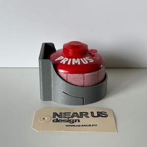 May include: A red Primus gas canister in a grey holder. The canister has white text and a red cap. The holder is a curved shape with a rectangular back. A tag with the text "NEAR US design" is in front.