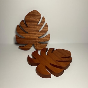 Puede incluir: Dos posavasos de madera con forma de hojas de monstera. Los posavasos son de color marrón oscuro y tienen una veta de madera natural.