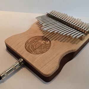 May include: A wooden kalimba instrument with a carved design and metal tines. The instrument has a light wood finish with a dark brown border. A silver-colored jack is connected to the instrument.