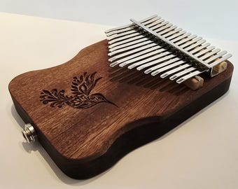 May include: A wooden kalimba with a hummingbird design etched on the surface. The instrument has a dark brown finish and a set of silver tines. A jack is visible on the side. The kalimba is a musical instrument.