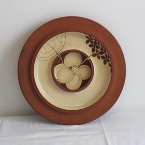 Puede incluir: Un conjunto de tres platos de cerámica en una disposición anidada. El plato más grande tiene un borde marrón rojizo, que enmarca un interior color crema con un diseño floral. El plato del medio es de color crema con un diseño floral, y el más pequeño es marrón rojizo.