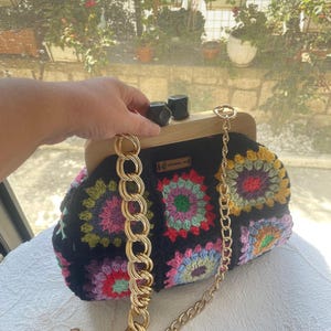 May include: A black crochet clutch with a wooden frame and a gold chain strap. The clutch features colorful floral patterns in pink, green, yellow, and blue. The wooden clasp is accented with two black square details.