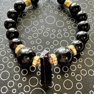 Può includere: Braccialetto con perline di onice nero con chiusura dorata e ciondolo di cristallo nero. Le perline sono accentate con oro e strass trasparenti.