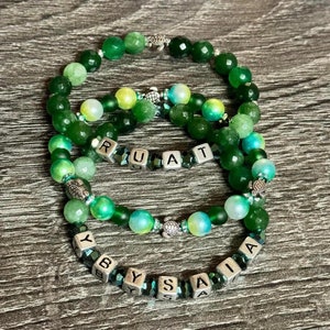 Puede incluir: Tres pulseras de cuentas verdes y azules con cuentas de amuleto plateadas que deletrean los nombres "RUAT" e "YBYSAIA".