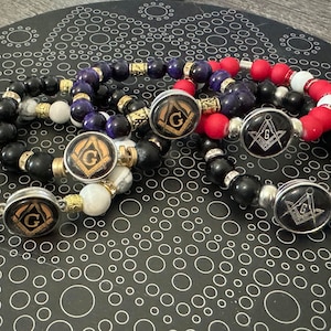 May include: Four bracelets with black, white, red, and blue beads. Each bracelet has a silver charm with a gold Masonic symbol.