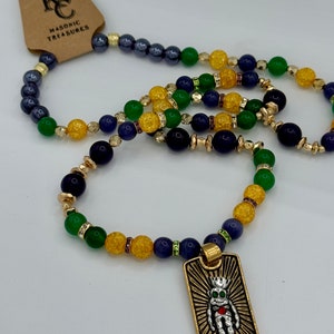 Puede incluir: Pulseras de cuentas con un colgante dorado que presenta una figura coronada con un corazón. Las pulseras tienen una mezcla de cuentas verdes, amarillas, azules y doradas. Una etiqueta marrón con el texto "Masonic Treasures" está adjunta.
