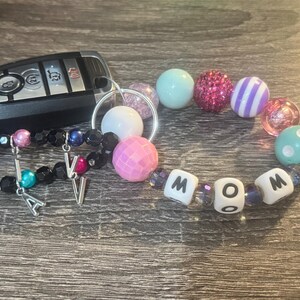 Pode incluir: Um chaveiro de contas coloridas com a palavra "MOM" escrita em contas brancas. O chaveiro tamb&eacute;m tem um anel de chave prateado e dois pingentes prateados com as letras "A" e "V".