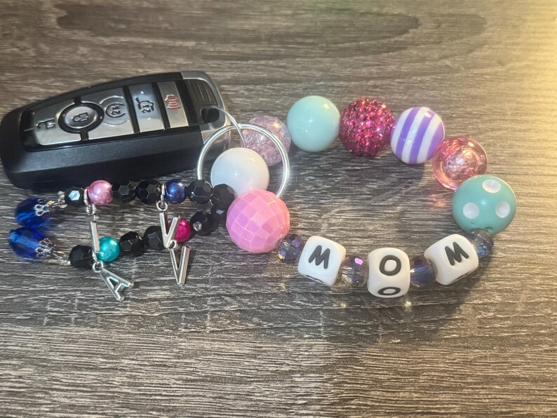 Pode incluir: Um chaveiro de contas coloridas com a palavra "MOM" escrita em contas brancas. O chaveiro tamb&eacute;m tem um anel de chave prateado e dois pingentes prateados com as letras "A" e "V".
