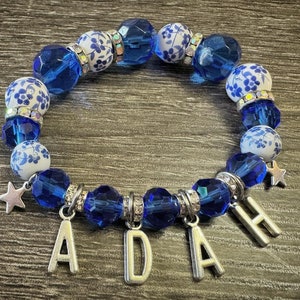 Puede incluir: Pulsera de cuentas azules y blancas con un dije de plata que dice "ADAH". Las cuentas son una mezcla de vidrio azul y cerámica blanca con un patrón floral azul.