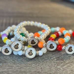 May include: Four colorful beaded bracelets with silver charms featuring a profile image of a woman wearing a helmet. The bracelets are layered on a wooden surface.