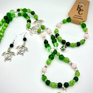 Puede incluir: Un conjunto de collar y pulsera de cuentas verdes y blancas con dijes de tortuga plateados. El conjunto incluye un par de aretes con dijes de tortuga plateados y cuentas negras. La joyería está etiquetada como "KC Masonic Treasures".