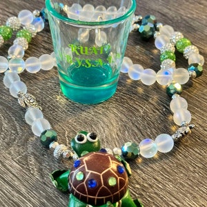 Puede incluir: Un collar de cuentas blancas y verdes con un dije de tortuga plateado y un vaso de chupito transparente con el texto "RUAT? BY.S.A." en verde.