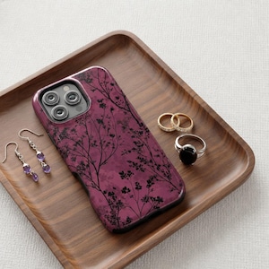 May include: A smartphone case with a deep magenta background featuring a black silhouette pattern of delicate tree branches and small leaves. The case is accompanied by a pair of dangling earrings with purple stones and three metal rings.