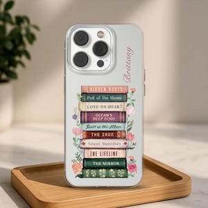 K&ouml;nnte beinhalten: Eine transparente Handyh&uuml;lle mit einem Stapel von B&uuml;chern, darunter Titel wie "Hidden Roots" und "The Lifeline." Die H&uuml;lle hat ein Blumenmuster und den Namen "Brittany." Die Handyh&uuml;lle liegt auf einem Holztablett.
