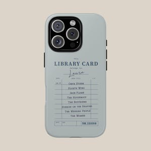 May include: A light gray phone case featuring a library card design. The text reads "This LIBRARY CARD belongs to: Laura" with a list of book titles. The case has a camera cutout and black accents on the sides.