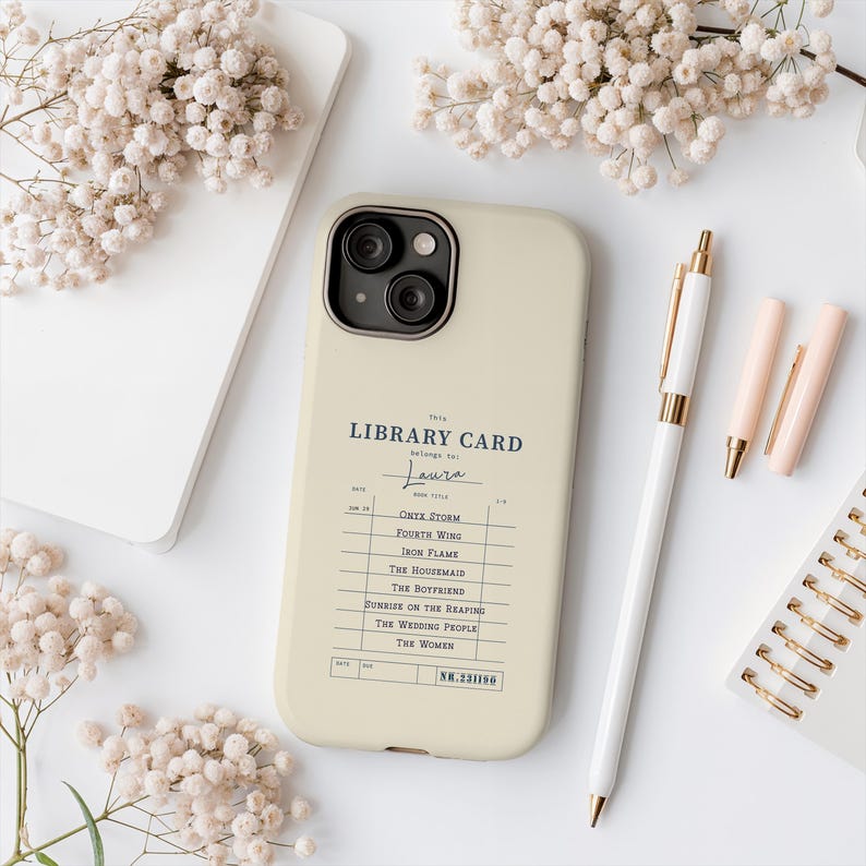 May include: A cream-colored phone case featuring a vintage library card design with the text "LIBRARY CARD" and a list of book titles. A white pen and two pink lipsticks are next to the phone case. The background includes white flowers and a white notebook.