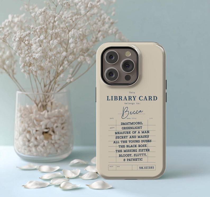 May include: A beige phone case featuring a library card design with the name "Becca" and a list of book titles. The case has a triple-lens camera cutout and a minimalist aesthetic. White flower petals and a vase of flowers are in the background.