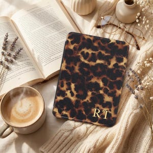 Op de afbeelding: Een tablet hoesje met luipaardprint en gouden initialen "RT" ligt op een crèmekleurige deken naast een open boek, een kopje koffie en een bril. De hoes heeft een bruin en beige vlekkenpatroon.