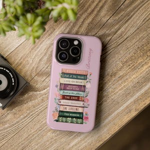 K&ouml;nnte beinhalten: Hellrosa Handyh&uuml;lle mit einem Stapel B&uuml;cher, darunter Titel wie "Hidden Roots" und "Love on Heart". Blumenschmuck und der Name "Brittany" sind ebenfalls zu sehen. Auf einer Holzoberfl&auml;che platziert.